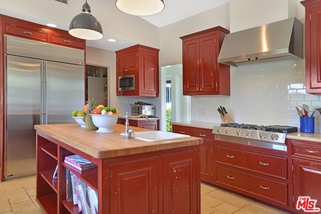 kitchen featuring built in appliances, a center island, butcher block counters, wall chimney range hood, and tasteful backsplash