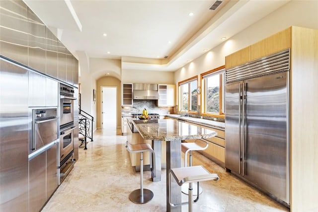 kitchen with wall chimney exhaust hood, a kitchen island, stone counters, appliances with stainless steel finishes, and a kitchen breakfast bar