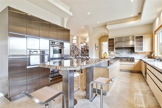 kitchen featuring light stone counters, a center island, stainless steel appliances, wall chimney range hood, and a kitchen breakfast bar