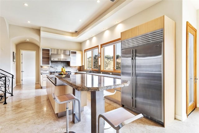 kitchen featuring wall chimney exhaust hood, built in refrigerator, tasteful backsplash, a kitchen island, and a breakfast bar