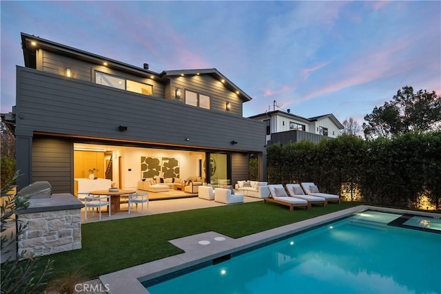 back of house at dusk with a balcony, a patio area, an outdoor hangout area, a pool with connected hot tub, and a lawn