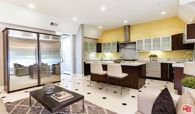 kitchen with dark brown cabinetry, vaulted ceiling, an island with sink, built in fridge, and wall chimney range hood