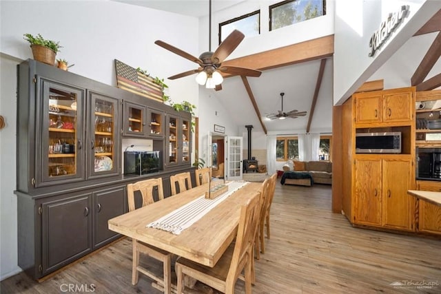 dining space featuring ceiling fan, a wood stove, light hardwood / wood-style floors, high vaulted ceiling, and beam ceiling