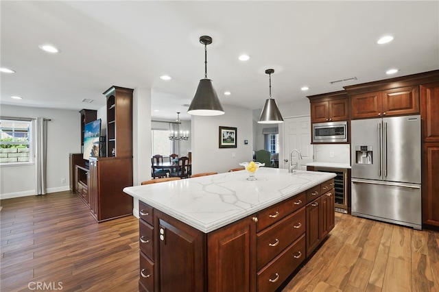 kitchen featuring light stone countertops, hardwood / wood-style floors, stainless steel appliances, hanging light fixtures, and a center island with sink