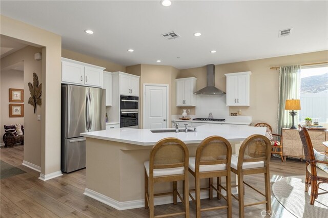 kitchen with a large island, white cabinetry, wall chimney exhaust hood, light wood-type flooring, and appliances with stainless steel finishes