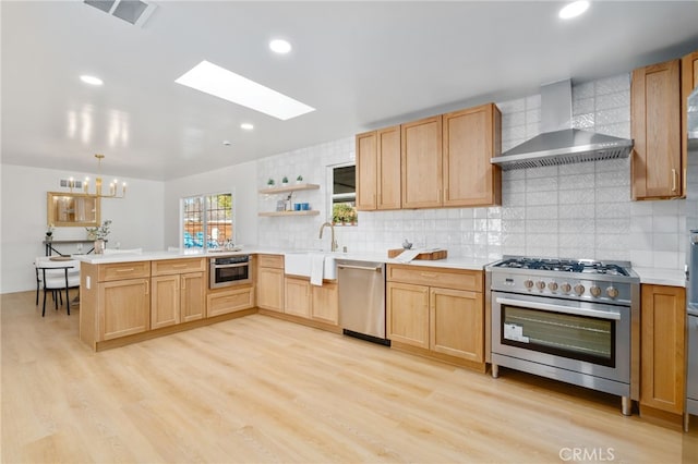 kitchen featuring appliances with stainless steel finishes, a skylight, kitchen peninsula, wall chimney exhaust hood, and light hardwood / wood-style flooring