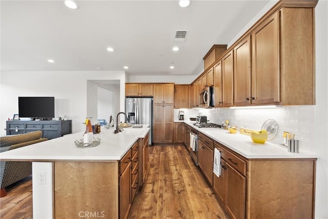 kitchen with an island with sink, appliances with stainless steel finishes, tasteful backsplash, light wood-type flooring, and sink