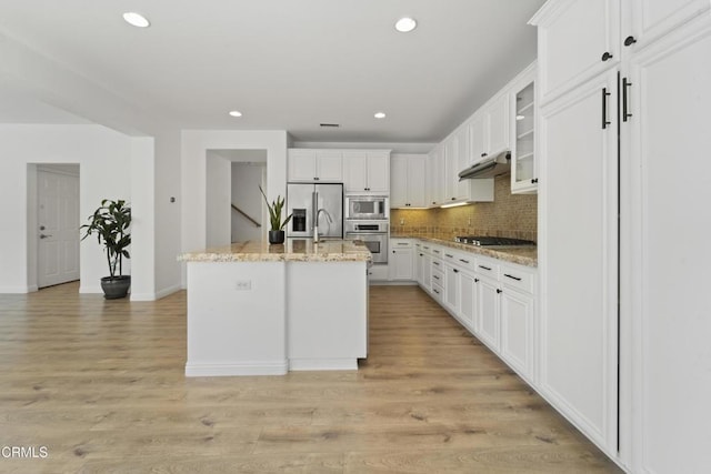 kitchen featuring a center island with sink, decorative backsplash, light stone countertops, stainless steel appliances, and white cabinets