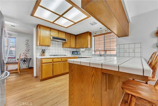 kitchen featuring tile countertops, kitchen peninsula, decorative backsplash, light wood-type flooring, and stainless steel appliances