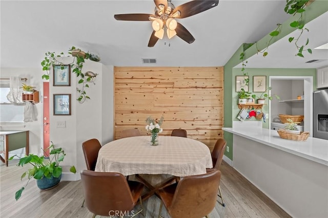 dining room featuring ceiling fan, light wood-type flooring, vaulted ceiling, and wood walls