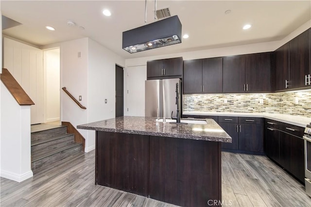 kitchen featuring dark brown cabinetry, stainless steel fridge, a kitchen island with sink, and dark stone counters