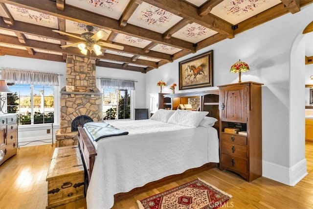 bedroom with beamed ceiling, a stone fireplace, light wood-type flooring, ceiling fan, and coffered ceiling