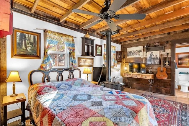 bedroom featuring ceiling fan, a wood stove, wood ceiling, connected bathroom, and beamed ceiling