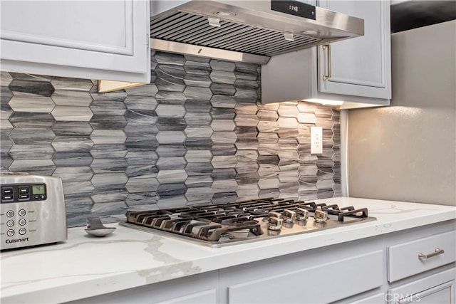 kitchen featuring stainless steel gas cooktop, light stone countertops, decorative backsplash, white cabinetry, and wall chimney range hood