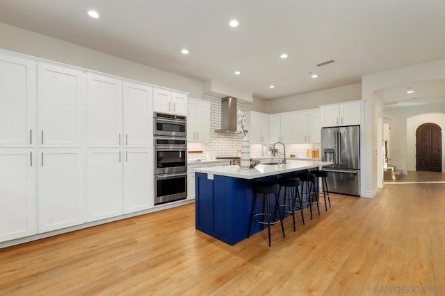 kitchen featuring stainless steel appliances, wall chimney range hood, a kitchen island with sink, white cabinets, and sink