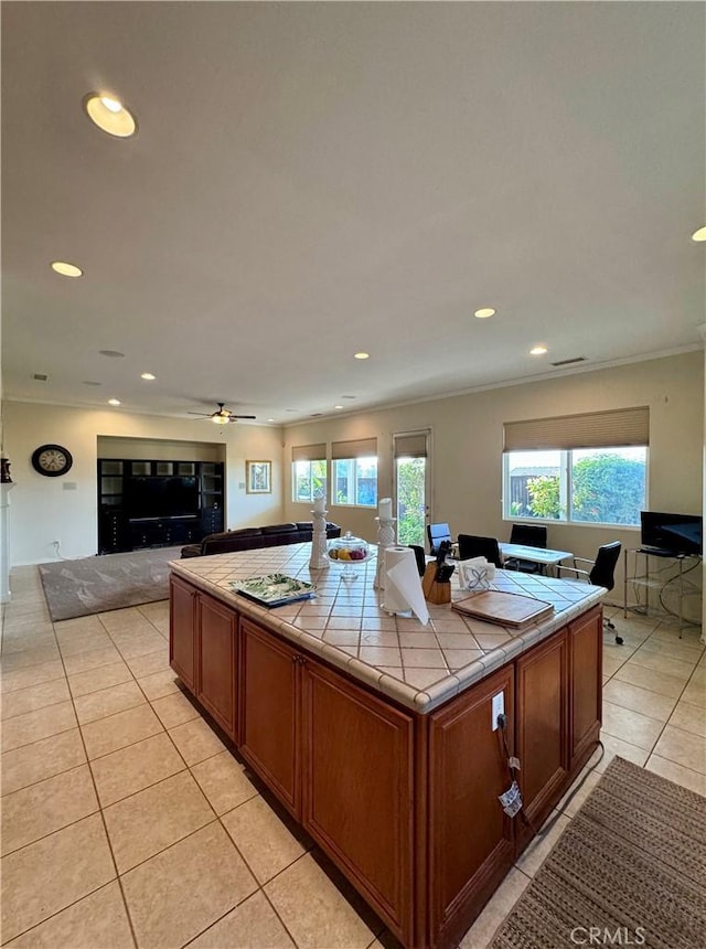 kitchen with light tile patterned flooring, ceiling fan, tile counters, a kitchen island, and crown molding