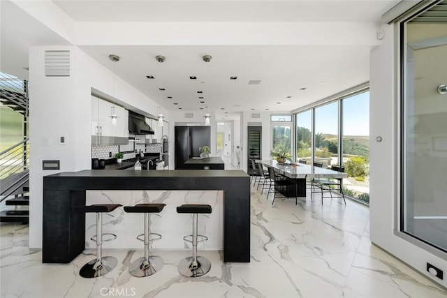 kitchen featuring black refrigerator, a breakfast bar area, wall chimney exhaust hood, white cabinets, and expansive windows