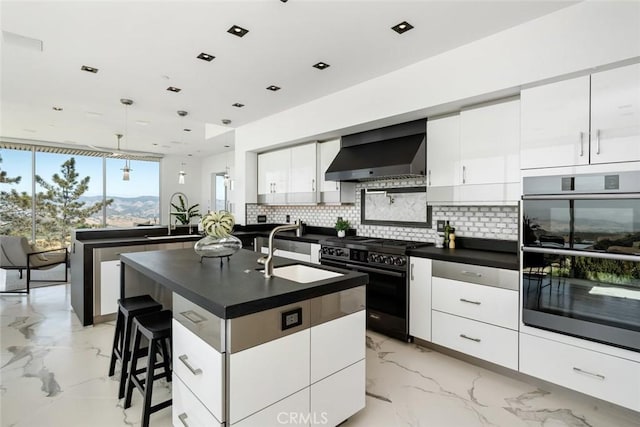 kitchen featuring wall chimney range hood, sink, black gas stove, a kitchen island with sink, and double oven