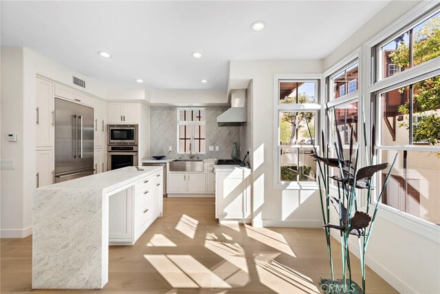 kitchen featuring sink, built in appliances, white cabinetry, wall chimney exhaust hood, and decorative backsplash