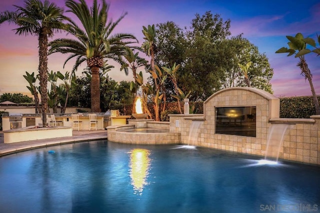 pool at dusk with pool water feature, exterior kitchen, and an in ground hot tub