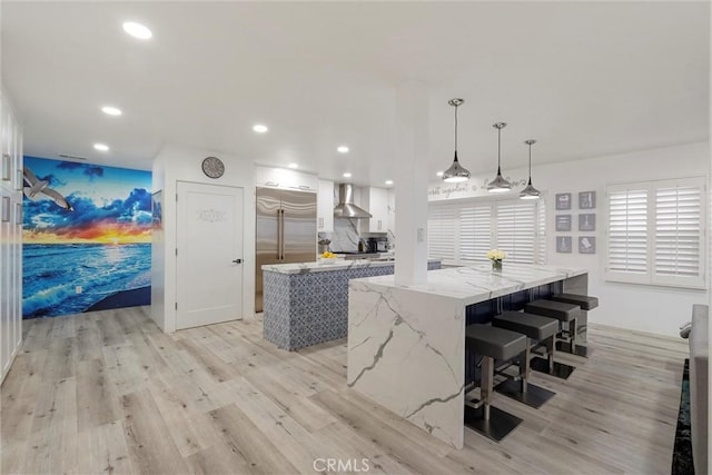 kitchen featuring light stone countertops, decorative light fixtures, wall chimney range hood, a kitchen island, and white cabinetry