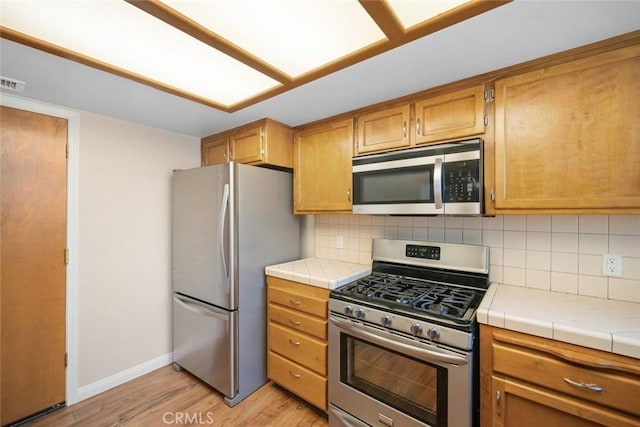 kitchen with light wood-type flooring, appliances with stainless steel finishes, tile counters, and tasteful backsplash