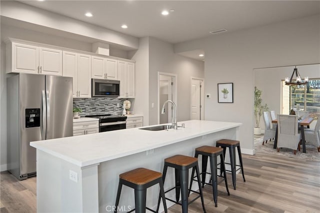 kitchen with stainless steel appliances, a notable chandelier, a kitchen island with sink, white cabinetry, and sink