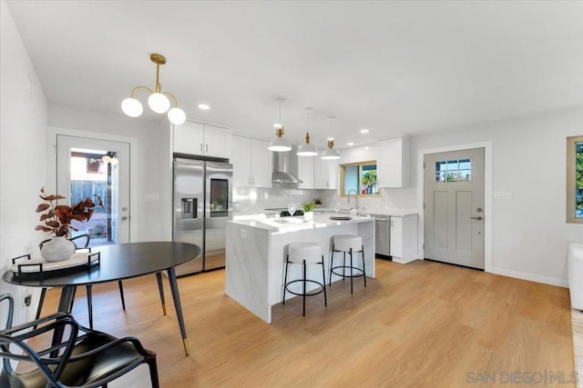 kitchen featuring hanging light fixtures, a center island, wall chimney range hood, stainless steel refrigerator with ice dispenser, and white cabinets