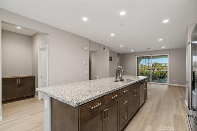 kitchen featuring sink, a kitchen island with sink, light stone countertops, stainless steel dishwasher, and light wood-type flooring