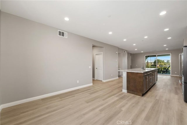 kitchen featuring dark brown cabinetry, light stone counters, a center island with sink, light hardwood / wood-style flooring, and stainless steel refrigerator