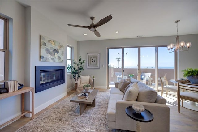 living room featuring recessed lighting, a glass covered fireplace, wood finished floors, baseboards, and ceiling fan with notable chandelier