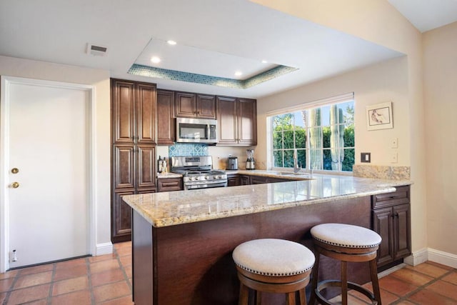 kitchen with a raised ceiling, appliances with stainless steel finishes, kitchen peninsula, light stone counters, and tasteful backsplash