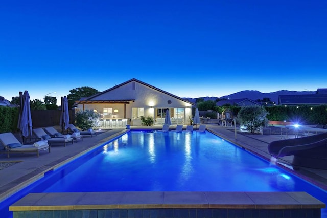 pool at dusk featuring a patio area, a water slide, a mountain view, and an outdoor structure