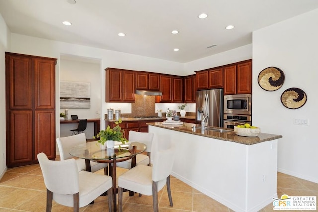 kitchen featuring light tile patterned floors, decorative backsplash, dark stone counters, a kitchen island with sink, and appliances with stainless steel finishes