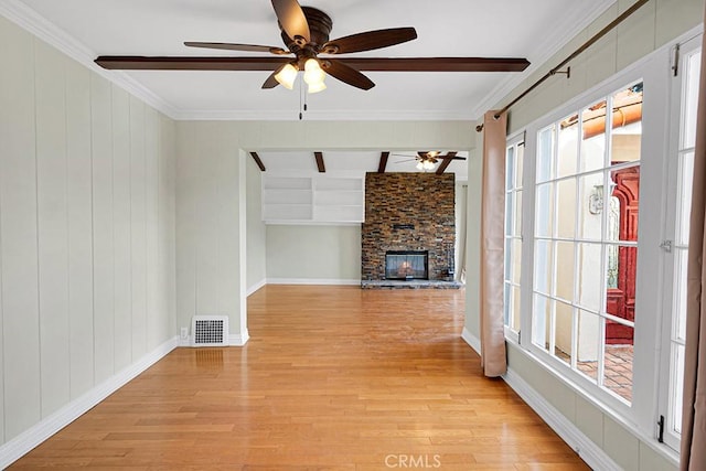 unfurnished living room featuring beam ceiling, a stone fireplace, ceiling fan, light hardwood / wood-style flooring, and crown molding