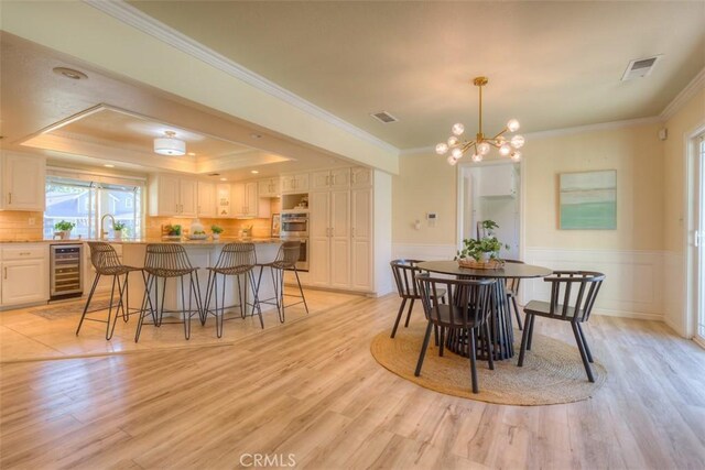 dining space with crown molding, an inviting chandelier, a tray ceiling, beverage cooler, and light wood-type flooring