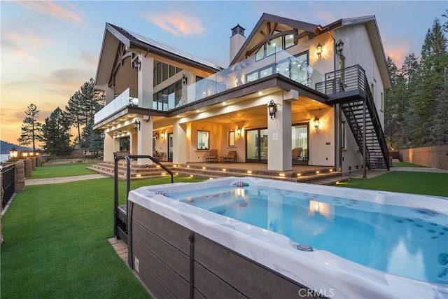 back house at dusk featuring a balcony, a patio area, a yard, and a hot tub