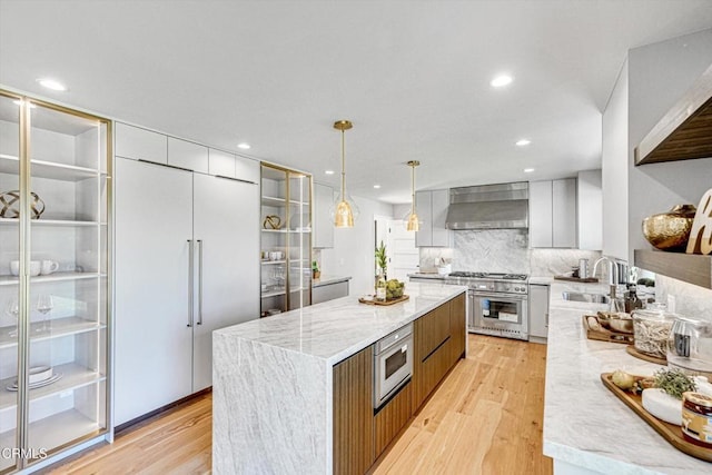 kitchen featuring hanging light fixtures, light stone countertops, wall chimney exhaust hood, built in appliances, and sink