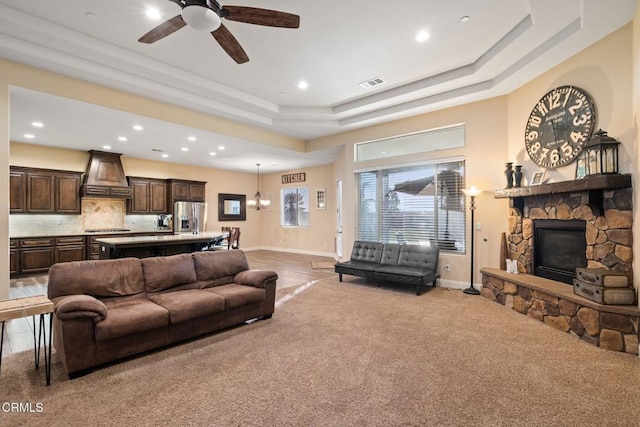 carpeted living room featuring ceiling fan, a stone fireplace, and a tray ceiling