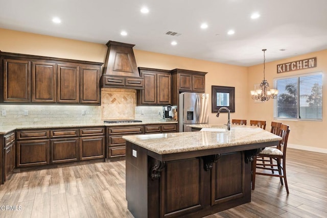 kitchen featuring a center island with sink, custom range hood, a notable chandelier, stainless steel fridge, and sink