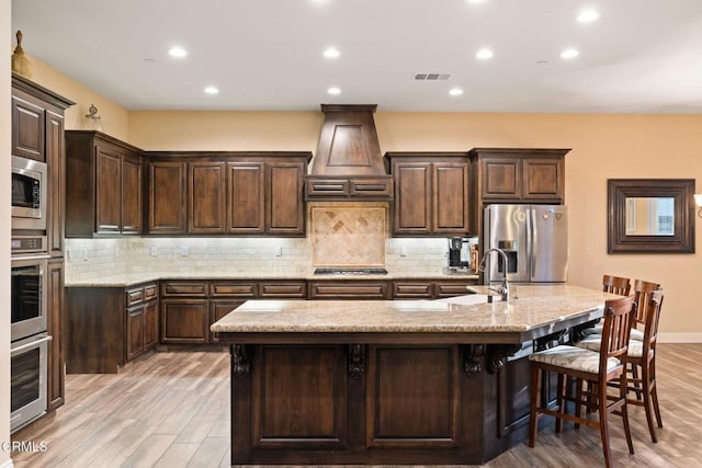 kitchen featuring stainless steel appliances, premium range hood, an island with sink, and a breakfast bar area