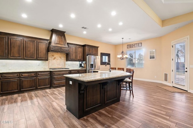 kitchen featuring decorative light fixtures, a kitchen bar, an island with sink, premium range hood, and appliances with stainless steel finishes