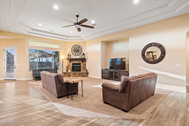 living room with ceiling fan, light hardwood / wood-style flooring, a stone fireplace, and a tray ceiling