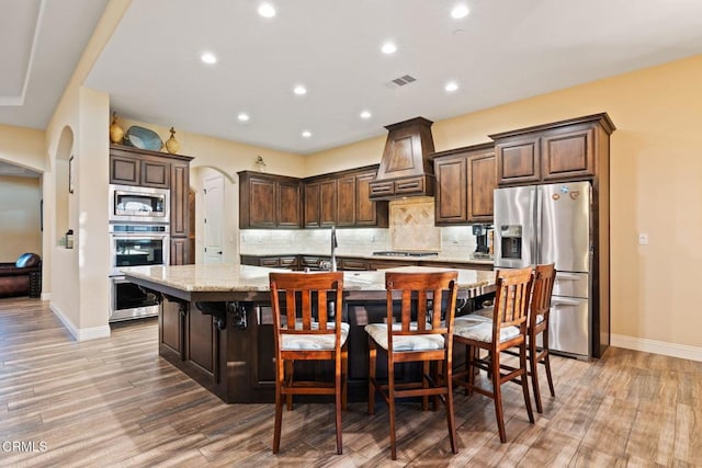 kitchen with appliances with stainless steel finishes, an island with sink, premium range hood, a breakfast bar, and light stone counters