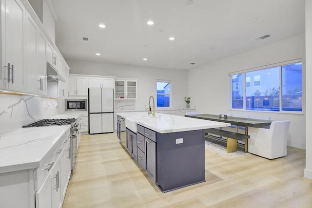 kitchen featuring light hardwood / wood-style floors, stainless steel appliances, a large island with sink, light stone counters, and white cabinets