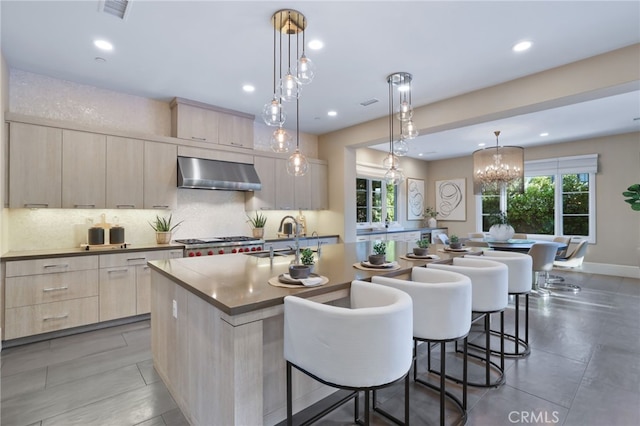 kitchen featuring an island with sink, pendant lighting, light brown cabinetry, sink, and wall chimney range hood