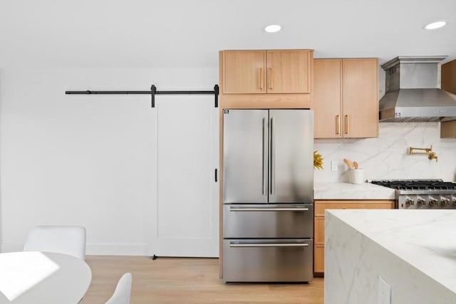 kitchen with light stone counters, decorative backsplash, a barn door, wall chimney range hood, and appliances with stainless steel finishes