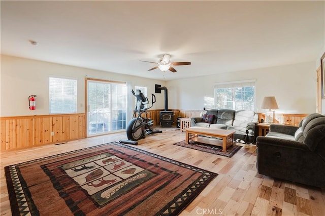 living room with a ceiling fan, a wood stove, a wainscoted wall, wooden walls, and light wood-style flooring