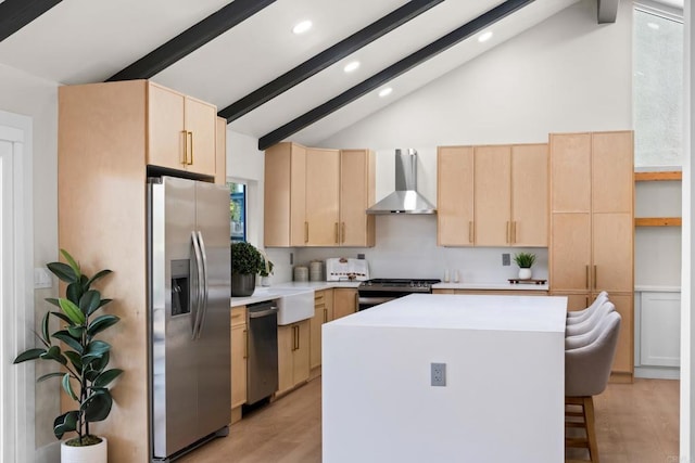 kitchen with appliances with stainless steel finishes, light wood-type flooring, a kitchen island, wall chimney exhaust hood, and beam ceiling