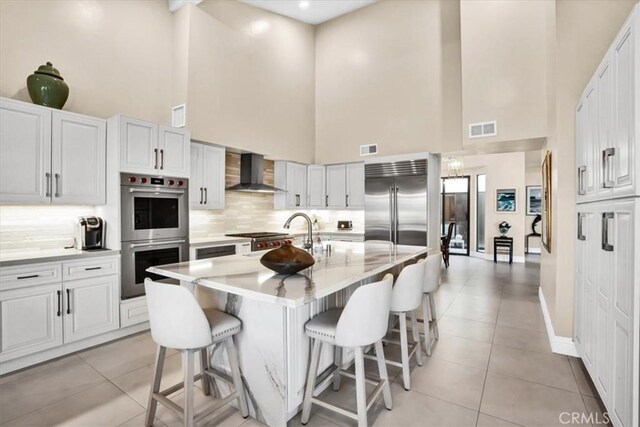 kitchen with white cabinets, a towering ceiling, a kitchen breakfast bar, wall chimney range hood, and appliances with stainless steel finishes